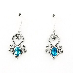 May include: A pair of silver-toned drop earrings. Each earring features a heart-shaped design with an oval, bright blue gemstone. The earrings have a decorative, ornate style with a hook closure.