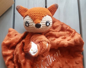 May include: A crocheted fox plush toy with large eyes and a brown and white color scheme. The fox is attached to a soft, orange, textured blanket with the name "Malhon" embroidered on it.