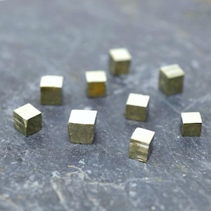 May include: A collection of small, metallic gold pyrite cubes. The cubes are scattered on a dark gray surface. Each cube has a distinct, geometric shape and a reflective, golden sheen, suggesting a natural mineral.