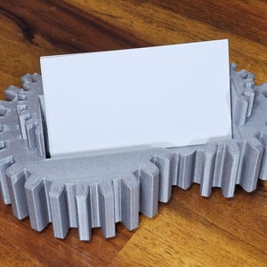 May include: A gray 3D printed business card holder shaped like a gear. The holder has a slot for a business card and is made of a textured plastic.