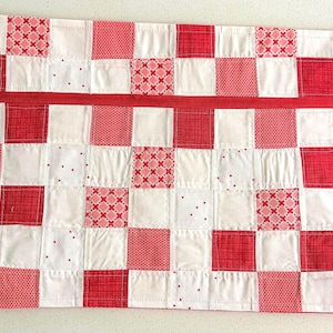 May include: A rectangular zippered pouch featuring a patchwork design of alternating white and red fabric squares. The red squares display various patterns including solid red, small dots, and cross-hatch motifs. A red zipper runs horizontally across the top.
