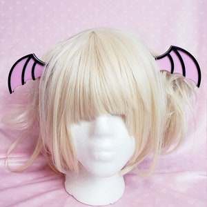 May include: A blonde wig styled with two pigtails, adorned with a pair of bat wing hair clips. The clips feature a black outline and pink interior, creating a striking contrast. The wig is displayed on a white foam head against a pink background.