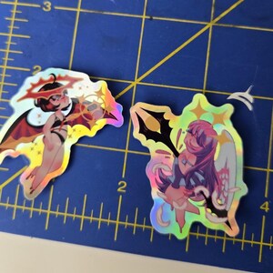 May include: Two holographic stickers featuring anime-style characters. One depicts a character with red wings and a halo, the other shows a character with white wings and a halo. Both stickers have a shiny, iridescent finish.