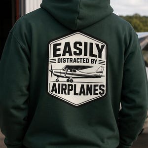 Może przedstawiać: Ciemnozielona bluza z kapturem z biało-czarną grafiką na plecach z napisem "EASILY DISTRACTED BY AIRPLANES" z ilustracją samolotu. Bluza z kapturem jest noszona przez osobę stojącą w pobliżu samolotu.