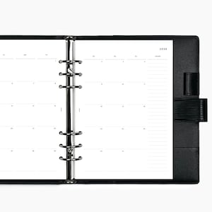May include: Open black planner with a monthly calendar layout for the year 2026. The planner features a silver metal ring binder and a black leather cover with a strap closure. The pages are white with black text.