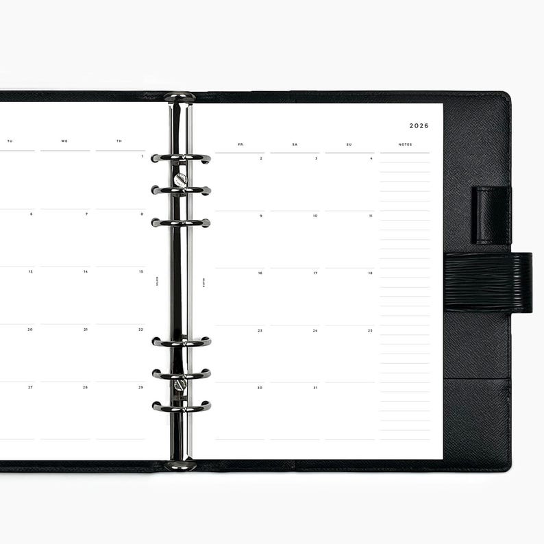 Puede incluir: Agenda negra abierta con un dise&ntilde;o de calendario mensual para el a&ntilde;o 2026. La agenda presenta una encuadernaci&oacute;n de anillas de metal plateado y una cubierta de cuero negro con cierre de correa. Las p&aacute;ginas son blancas con texto negro.