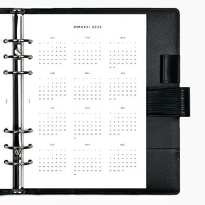 May include: A black leather binder with a 2026 calendar insert. The calendar pages are white with black text, displaying the months and dates. The binder has a silver metal ring mechanism and a black strap closure.