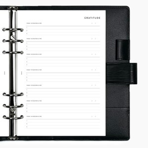 May include: A black leather-bound planner with a white page open to a gratitude journal section. The page has lines for writing and the heading "GRATITUDE".