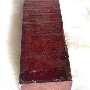 May include: A long, rectangular block of dark reddish-brown wood with a glossy, polished finish. The surface displays a visible grain pattern and texture, with slightly rough, unfinished edges along the sides and a flat, square end face.