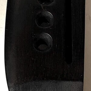 K&ouml;nnte beinhalten: Eine schwarze Gitarrenbr&uuml;cke aus Holz mit sechs Saitenl&ouml;chern und einem vertikalen Schlitz. Die Br&uuml;cke ist etwa 9 cm lang und befindet sich neben einem gelben Ma&szlig;band.