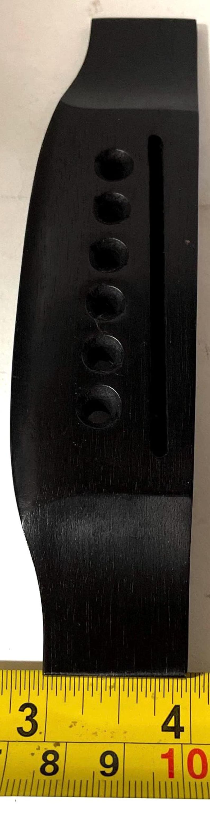 K&ouml;nnte beinhalten: Eine schwarze Gitarrenbr&uuml;cke aus Holz mit sechs Saitenl&ouml;chern und einem vertikalen Schlitz. Die Br&uuml;cke ist etwa 9 cm lang und befindet sich neben einem gelben Ma&szlig;band.