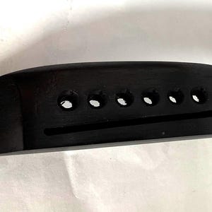 K&ouml;nnte beinhalten: Eine schwarze Gitarrenbr&uuml;cke mit sechs Saitenl&ouml;chern und einem Schlitz f&uuml;r den Steg. Die Br&uuml;cke besteht aus einem dunklen, soliden Material und hat eine glatte, polierte Oberfl&auml;che. Dieses Gitarrenteil dient dazu, die Saiten zu halten.