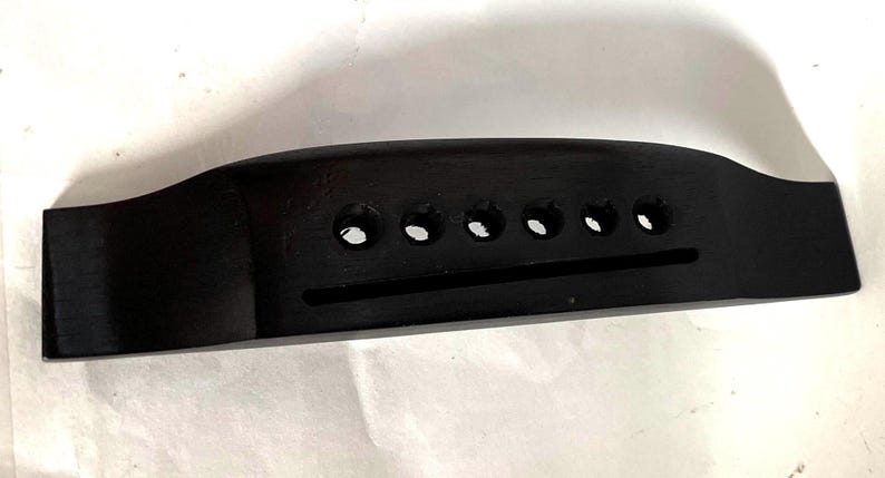 K&ouml;nnte beinhalten: Eine schwarze Gitarrenbr&uuml;cke mit sechs Saitenl&ouml;chern und einem Schlitz f&uuml;r den Steg. Die Br&uuml;cke besteht aus einem dunklen, soliden Material und hat eine glatte, polierte Oberfl&auml;che. Dieses Gitarrenteil dient dazu, die Saiten zu halten.