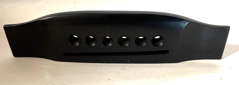 K&ouml;nnte beinhalten: Eine schwarze Gitarrenbr&uuml;cke mit sechs Saitenl&ouml;chern und einem Schlitz f&uuml;r den Steg. Die Br&uuml;cke ist aus Holz gefertigt und hat eine glatte, gl&auml;nzende Oberfl&auml;che. Sie dient dazu, die Saiten auf einer Akustikgitarre zu halten.