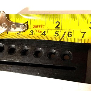 K&ouml;nnte beinhalten: Ein schwarzer Gitarrensteg mit sechs L&ouml;chern, gemessen mit einem gelben Ma&szlig;band. Das Ma&szlig;band zeigt ein Ma&szlig; von etwa 5 cm an.