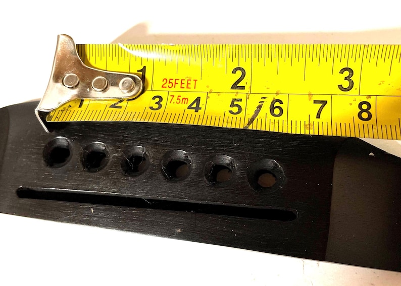 K&ouml;nnte beinhalten: Ein schwarzer Gitarrensteg mit sechs L&ouml;chern, gemessen mit einem gelben Ma&szlig;band. Das Ma&szlig;band zeigt ein Ma&szlig; von etwa 5 cm an.