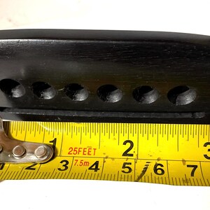 K&ouml;nnte beinhalten: Eine schwarze Gitarrenbr&uuml;cke aus Holz mit sechs Saitenl&ouml;chern und einem Schlitz f&uuml;r den Steg. Ein gelbes Ma&szlig;band zeigt, dass die Br&uuml;cke etwa 7,6 cm lang ist.