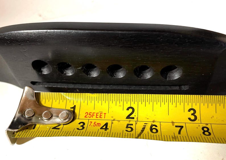 K&ouml;nnte beinhalten: Eine schwarze Gitarrenbr&uuml;cke aus Holz mit sechs Saitenl&ouml;chern und einem Schlitz f&uuml;r den Steg. Ein gelbes Ma&szlig;band zeigt, dass die Br&uuml;cke etwa 7,6 cm lang ist.