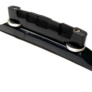 May include: A black wooden guitar bridge with a slotted top and two silver adjustment wheels. The bridge is designed to hold the strings in place and is a component of a stringed instrument.