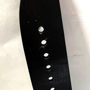 K&ouml;nnte beinhalten: Eine schwarze Gitarrenbr&uuml;cke mit sechs runden L&ouml;chern f&uuml;r Saiten. Die Br&uuml;cke ist aus Holz und hat eine gebogene Form. Der Hintergrund ist wei&szlig;.