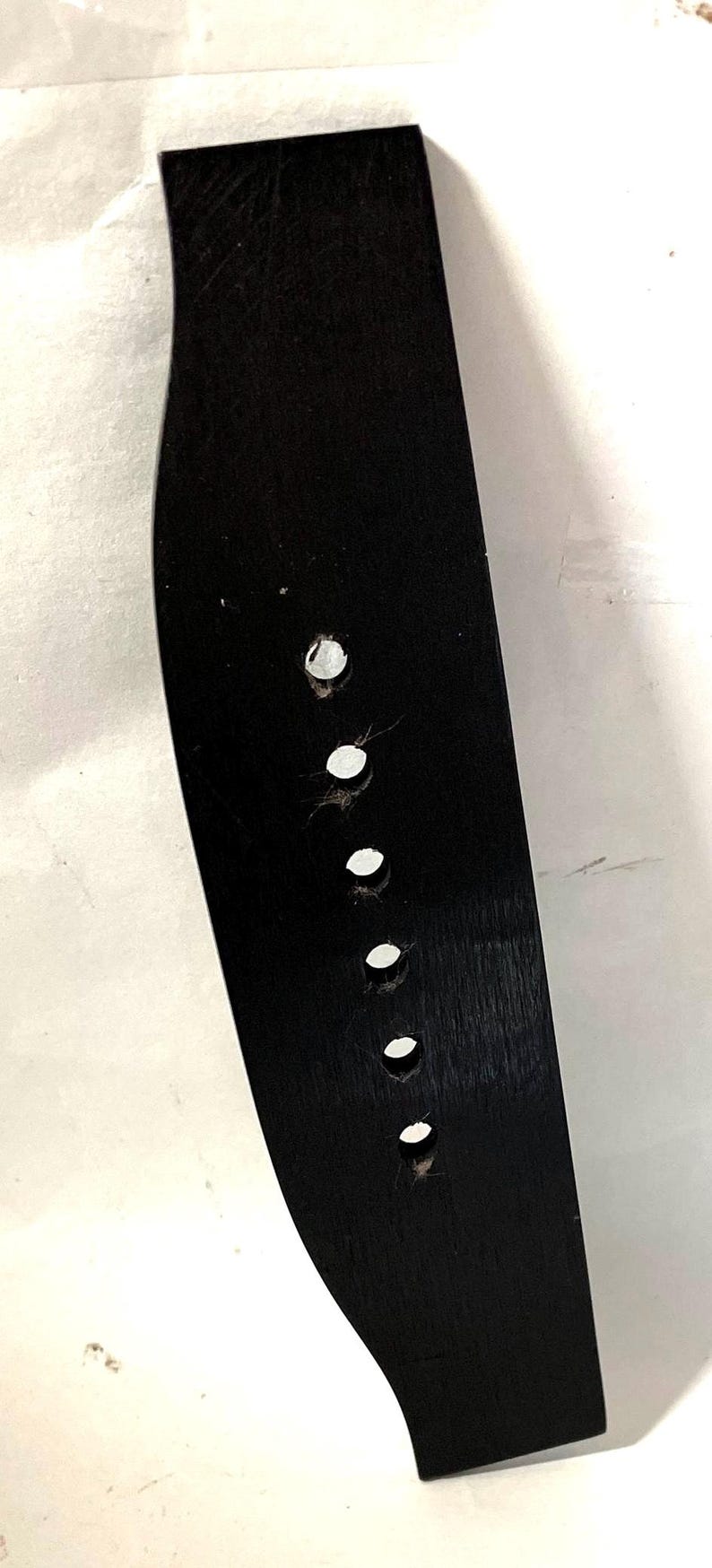 K&ouml;nnte beinhalten: Eine schwarze Gitarrenbr&uuml;cke mit sechs runden L&ouml;chern f&uuml;r Saiten. Die Br&uuml;cke ist aus Holz und hat eine gebogene Form. Der Hintergrund ist wei&szlig;.