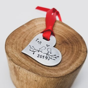 May include: Silver heart-shaped ornament with a red ribbon. The ornament is engraved with two birds on a branch, the initials "S+M", and the year "2019".