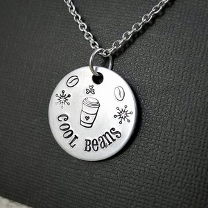 May include: A silver-colored pendant necklace with a round charm. The charm has the words "Cool Beans" and coffee-themed illustrations. The necklace is displayed on a dark textured surface.