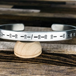 May include: Silver bracelet with a repeating pattern of ribbon symbols and a single slash symbol.