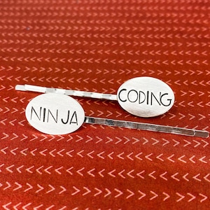 May include: Two silver hair clips with oval-shaped charms. The charms have the words "CODING" and "NINJA" engraved on them.