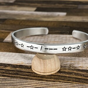 May include: Silver bangle bracelet with a pattern of stars and dashes. The bracelet is on a wooden surface.