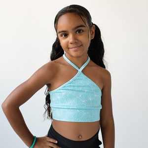 May include: A young person wearing a blue halter top with a white floral pattern and black athletic shorts. They are standing in front of a white background.