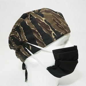 May include: A camouflage patterned surgical scrub hat with a black face mask and white ties. The hat has a button on the top.
