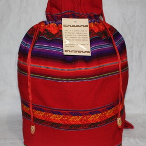 Puede incluir: Una bolsa roja con cordón y un patrón tejido colorido. La bolsa presenta un diseño vibrante con rayas horizontales en rojo, morado y azul. Una etiqueta con texto está adjunta. La bolsa está hecha de un material suave.