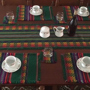 May include: A dining table set for a meal, featuring a dark brown table with a green and patterned table runner and placemats. White teacups and saucers, glasses, and a bottle are arranged on the table, creating a warm and inviting atmosphere.