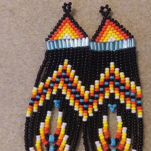 May include: A pair of long, beaded earrings with a black, orange, yellow, white, and blue geometric pattern. The earrings are made with small, round beads and have a fringe-like design.