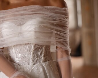 Puede incluir: Un vestido blanco de hombros descubiertos con un corpiño estilo corsé con intrincados bordados blancos y lentejuelas. El vestido incluye una capa de tul transparente y brillante drapeada sobre los hombros y guantes de tul a juego.