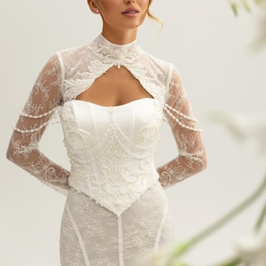 Könnte beinhalten: Weißes Brautkleid mit einem Spitzenbolero mit hohem Kragen, langen Ärmeln und Schlüssellochdetail. Das Oberteil ist mit aufwendigen Perlenarbeiten und einem Bustier aus Satin verziert. Der Rock ist ein eng anliegendes Spitzendesign. Ein klassisches Hochzeitskleid.