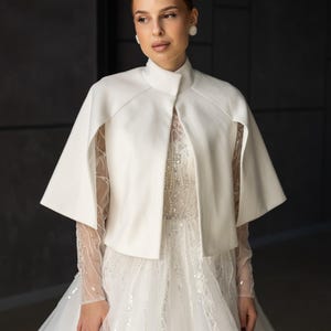 May include: A white bridal cape with a high collar and short sleeves. The cape is worn over a white wedding dress with long, sheer sleeves and intricate beading. The overall style is elegant and formal.