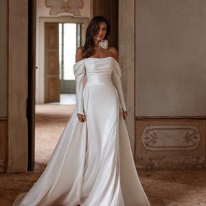Könnte beinhalten: Ein bodenlanges weißes Kleid mit schulterfreiem Ausschnitt, langen Ärmeln, einem figurbetonten Oberteil mit dezenten horizontalen Falten und einem fließenden Rock mit angesetztem Überrock. Das Kleid ist mit einer großen weißen Stoffblume verziert.