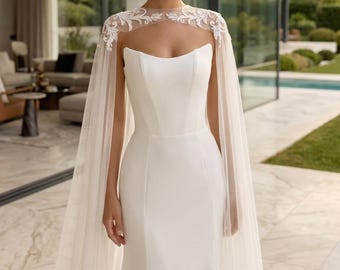Puede incluir: Un vestido blanco largo hasta el suelo con un corpiño ajustado de escote recto y una capa transparente. La capa está adornada con intrincados bordados florales blancos en los hombros y la parte superior del pecho, extendiéndose en largos paneles fluidos.