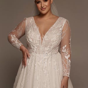 Puede incluir: Un vestido de novia blanco con escote pronunciado y mangas largas y transparentes. El vestido está adornado con intrincados bordados florales y lentejuelas brillantes. Se lleva un velo.
