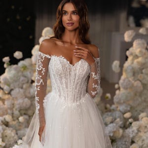 May include: An off-the-shoulder white wedding dress featuring a corset-style bodice with vertical boning, long sheer sleeves with intricate floral lace embroidery, and a voluminous tulle skirt adorned with subtle sparkling embellishments.