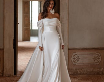 May include: A floor-length white gown featuring an off-the-shoulder neckline, long sleeves with buttoned cuffs, a ruched bodice, and a detachable overskirt. The dress is made of a smooth, satin-like fabric and includes a large fabric flower accent.