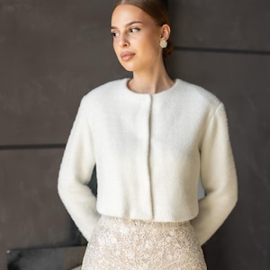 Puede incluir: Una mujer con una chaqueta corta blanca y difusa con cuello redondo y cierre de botón. También lleva una falda beige adornada. El fondo es una pared gris oscuro.