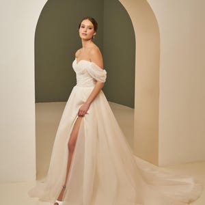 Op de afbeelding: Een ivoorkleurige trouwjurk met blote schouders, een hartvormige halslijn, een geplooid lijfje en een volumineuze tule rok met een hoge split. De japon tot op de grond heeft een lange sleep en fijne gedrapeerde mouwen over de schouders.