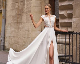 May include: A white floor-length gown featuring a sleeveless bodice with intricate white floral embroidery, a sheer illusion neckline, a lace-up corset-style midsection, and a long flowing skirt with a high side slit.