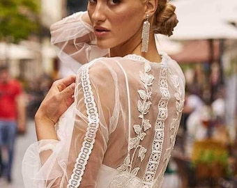 Könnte beinhalten: Ein weißes Kleid mit langen Ärmeln aus transparentem Tüll, aufwendigen weißen Blumenstickereien und Spitzenbesatz an den Armen und am Rücken. Das Kleidungsstück hat ein eng anliegendes Oberteil mit betonter Taille und einen fließenden Rock.