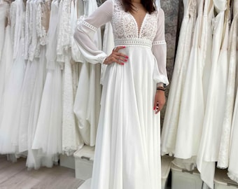 May include: A floor-length white gown featuring a deep V-neckline with intricate lace detailing on the bodice, long sheer sleeves with lace cuffs, and a flowing skirt. The waist is accented with a horizontal lace trim band.