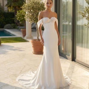 Puede incluir: Un vestido blanco largo hasta el suelo con escote de hombros caídos, un corpiño adornado con encaje y una estructura tipo corsé. La falda es ajustada en las caderas y se ensancha ligeramente hacia el suelo, con un dobladillo de encaje.