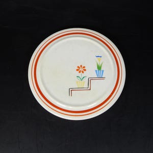 May include: A white ceramic plate with two concentric orange stripes around the rim. The plate features a floral design with orange, yellow, blue, and green flowers in pots, and a brown staircase design.